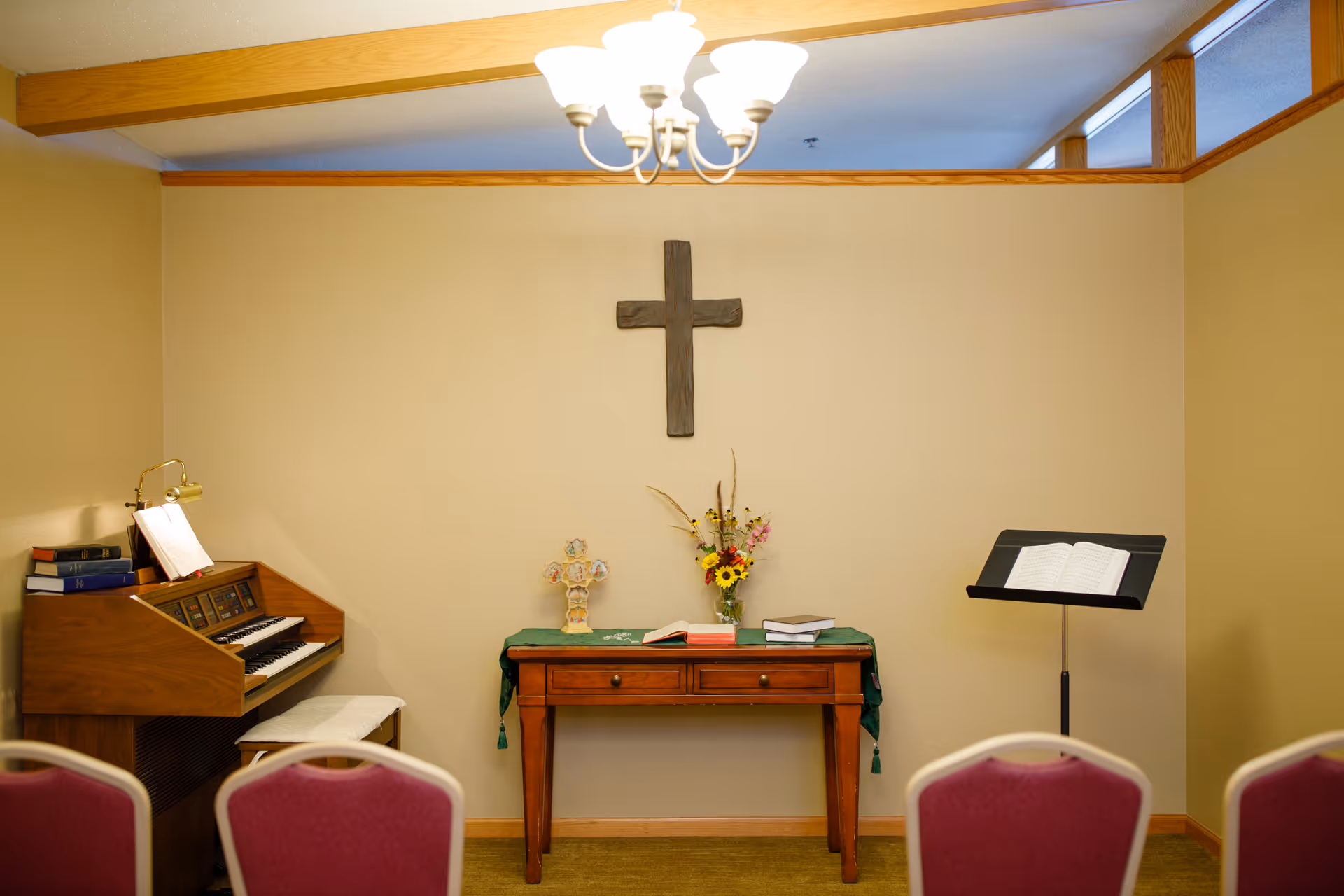 Small chapel room with a wooden cross mounted on a beige wall above a wooden table covered with a green cloth, flowers, and books. To the left is an organ with sheet music and a bench, and to the right is a music stand with an open book. Several red chairs are visible in the foreground.