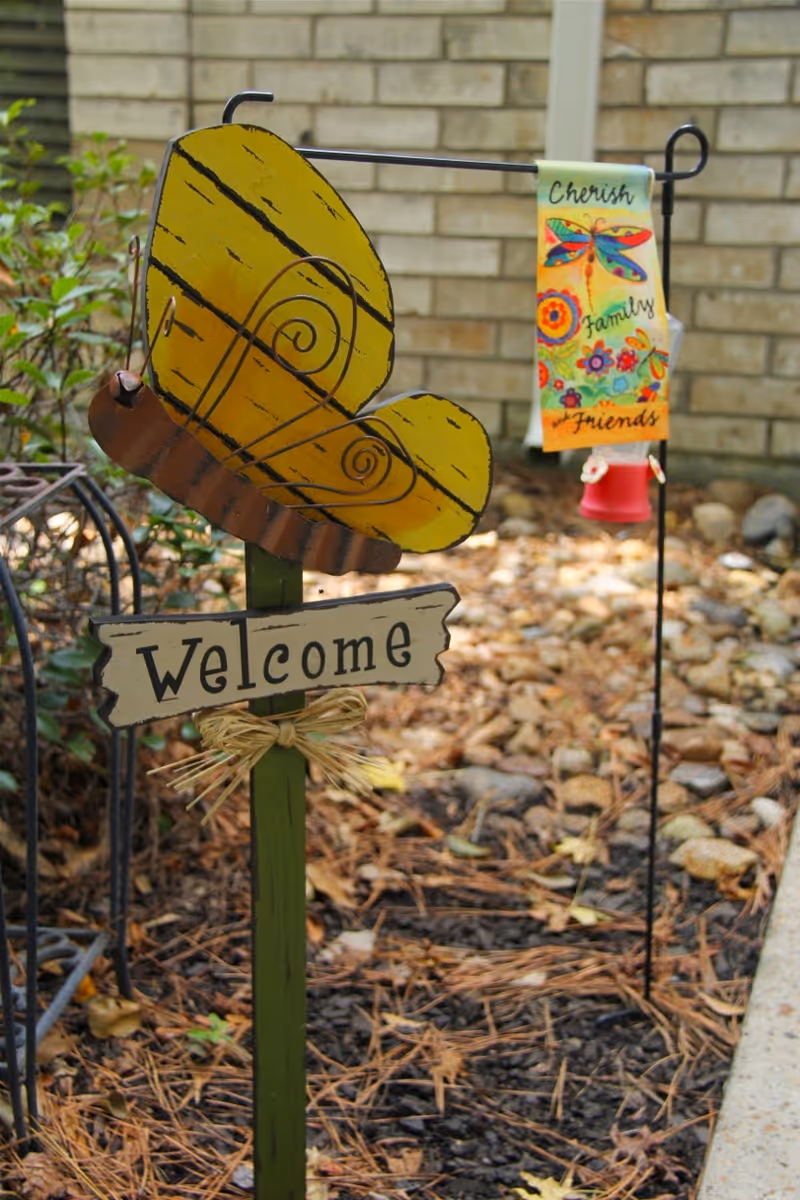 A garden decoration featuring a yellow butterfly with a rustic 'Welcome' sign attached to a green stake in the ground. In the background, there is a small colorful flag with a dragonfly and the words 'Cherish Family and Friends' hanging on a metal stand. The setting appears to be an outdoor garden area with rocks, plants, and a brick wall.