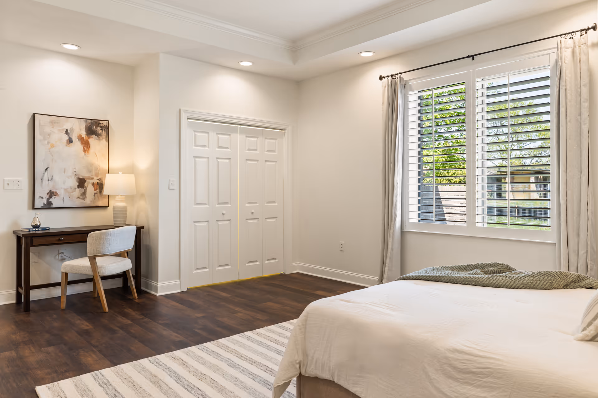 A bright bedroom with a large window featuring white plantation shutters and light curtains. The room has a bed with white bedding and a green throw blanket. There is a wooden desk with a beige upholstered chair, a table lamp, and abstract artwork on the wall. The floor is dark wood with a light striped rug.