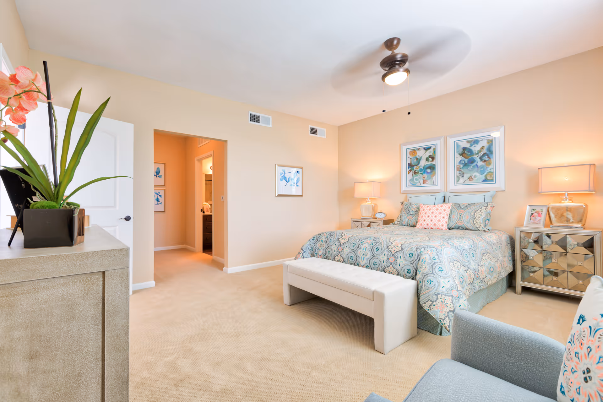 A bright and cozy bedroom with a large bed covered in a patterned blue and beige comforter, two matching bedside tables with lamps, a cushioned bench at the foot of the bed, a ceiling fan with light, and a light blue armchair with a decorative pillow. The walls are painted beige, and there are framed artworks above the bed and on the wall. A doorway leads to another room, possibly a bathroom.