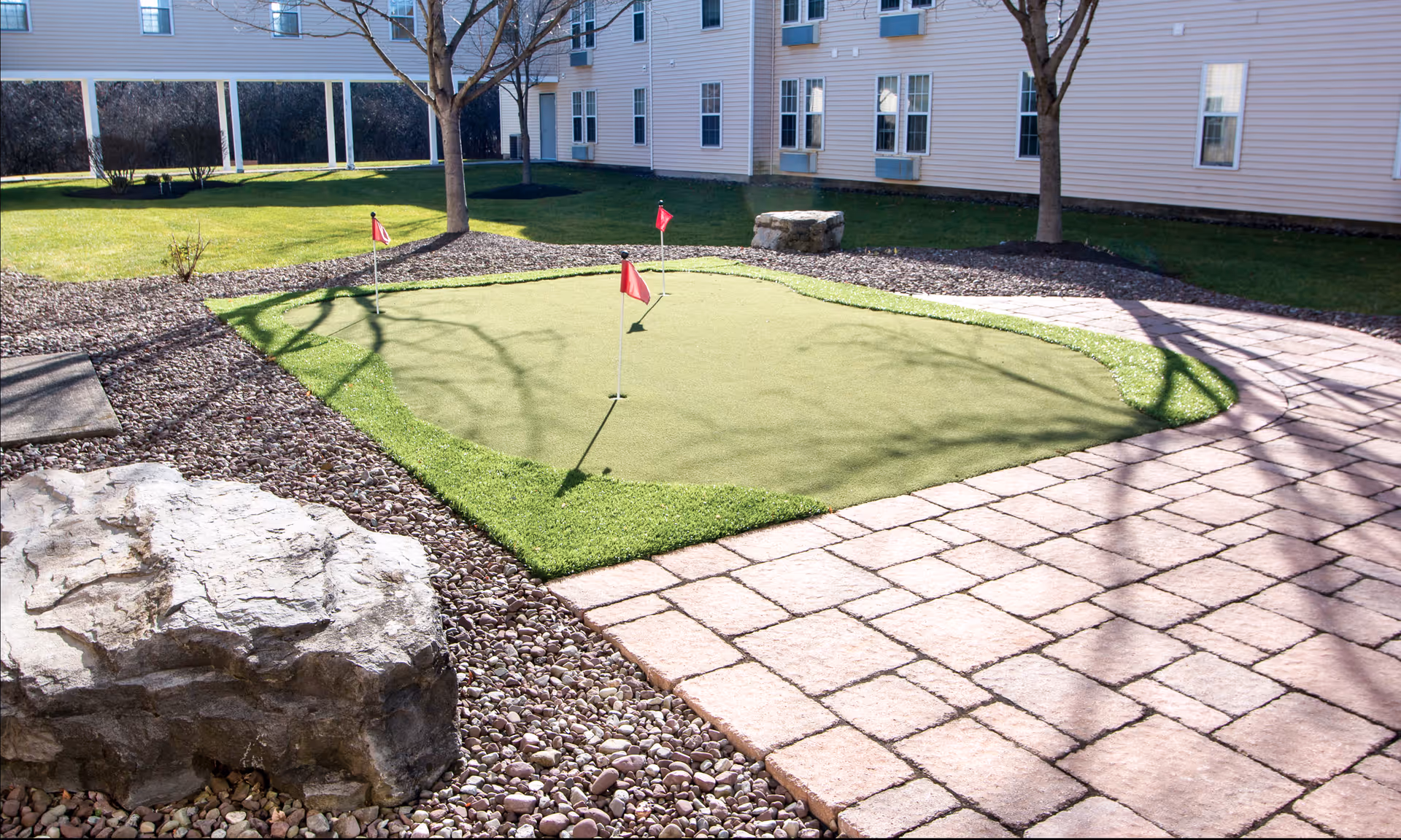A small outdoor courtyard putting green with three red flags, paved patio, decorative rocks, trees, and the exterior of a multi-story building.