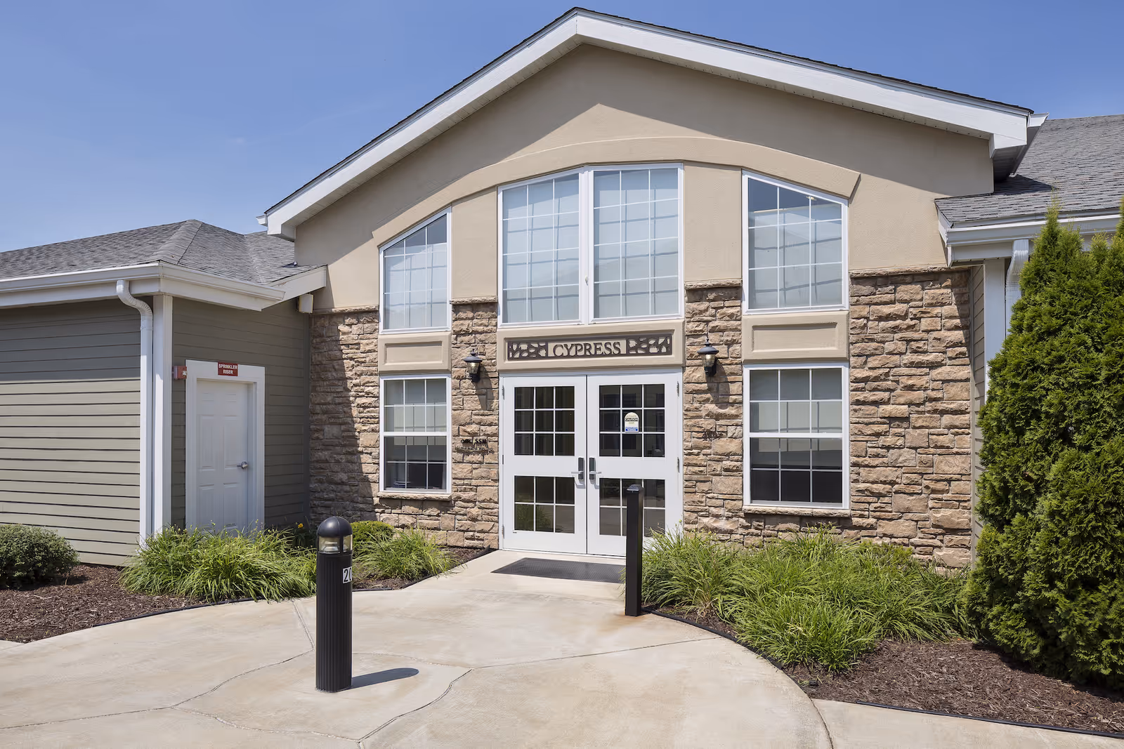 Exterior view of a senior living facility entrance with stone and beige siding, large windows, and double glass doors under a sign that reads 'CYPRESS'. There are plants and shrubs along the walkway leading to the entrance.