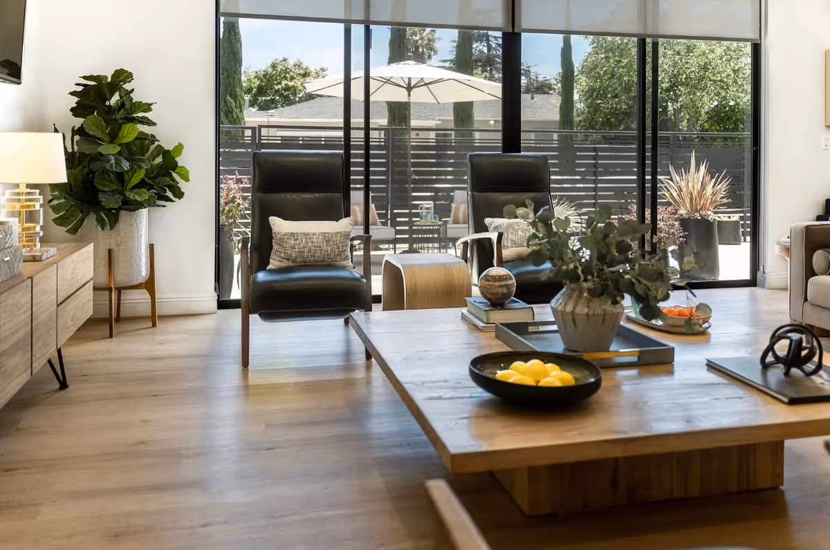 A modern living room with two black leather armchairs with pillows, a wooden coffee table with decorative items including a bowl of lemons, books, and a plant. Large floor-to-ceiling windows reveal an outdoor patio with an umbrella and greenery. A wooden sideboard with a lamp and a large potted plant is on the left side of the room.