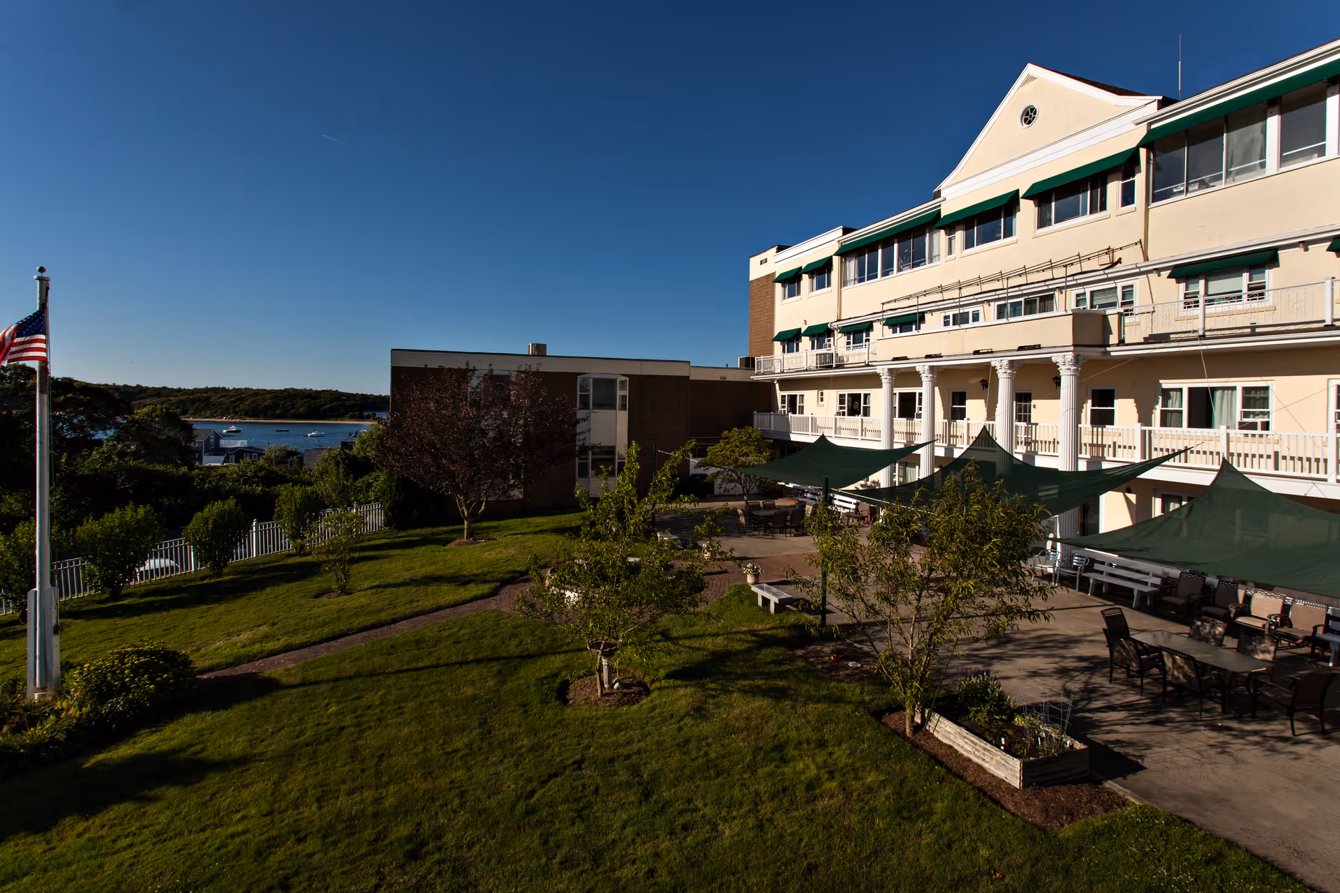 Outdoor view of Royal Megansett Nursing Home showing a multi-story building with balconies and green awnings. There is a well-maintained garden with trees, shrubs, and a paved patio area with tables and chairs shaded by green canopies. An American flag is visible on a flagpole to the left, with a body of water and boats in the background under a clear blue sky.