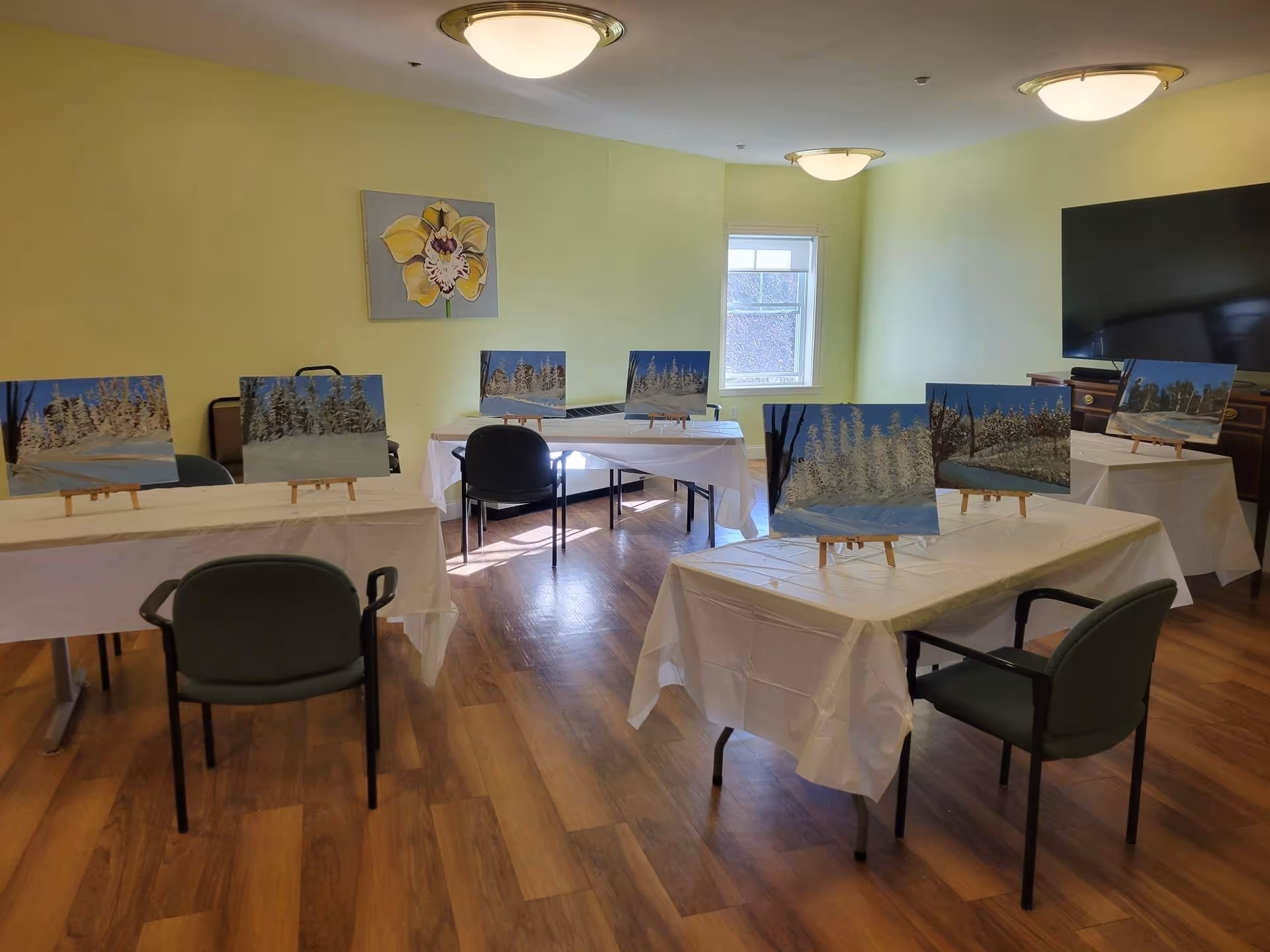 A well-lit room with light green walls and wooden flooring, featuring several tables covered with white tablecloths. Each table has small easels displaying paintings of snowy winter landscapes. There are green chairs placed around the tables, a window letting in natural light, a large flat-screen TV mounted on the wall, and a floral painting hanging on the wall.