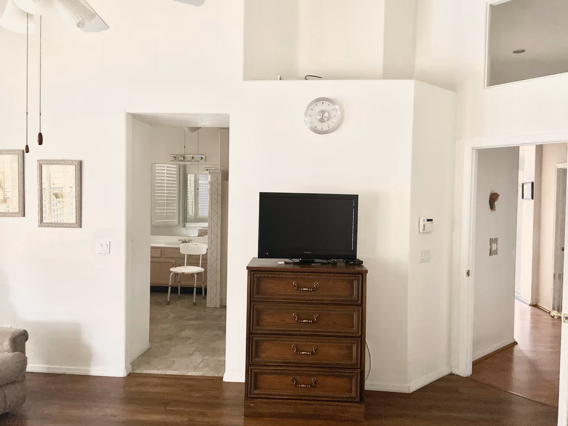 Interior view of a living space in an assisted living facility showing a wooden chest of drawers with a flat-screen TV on top, a wall clock above it, and an open doorway leading to a bathroom with a white chair and a sink visible. The room has white walls, wooden flooring, and a ceiling fan with pull chains.