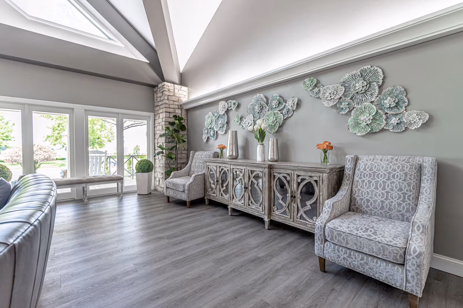 A bright and spacious living room area with high vaulted ceilings and large windows letting in natural light. The room features a gray leather sofa, two patterned armchairs, a decorative wooden sideboard with mirrored doors, and wall art consisting of floral sculptures. There are also potted plants and a bench near the windows overlooking a green outdoor area.
