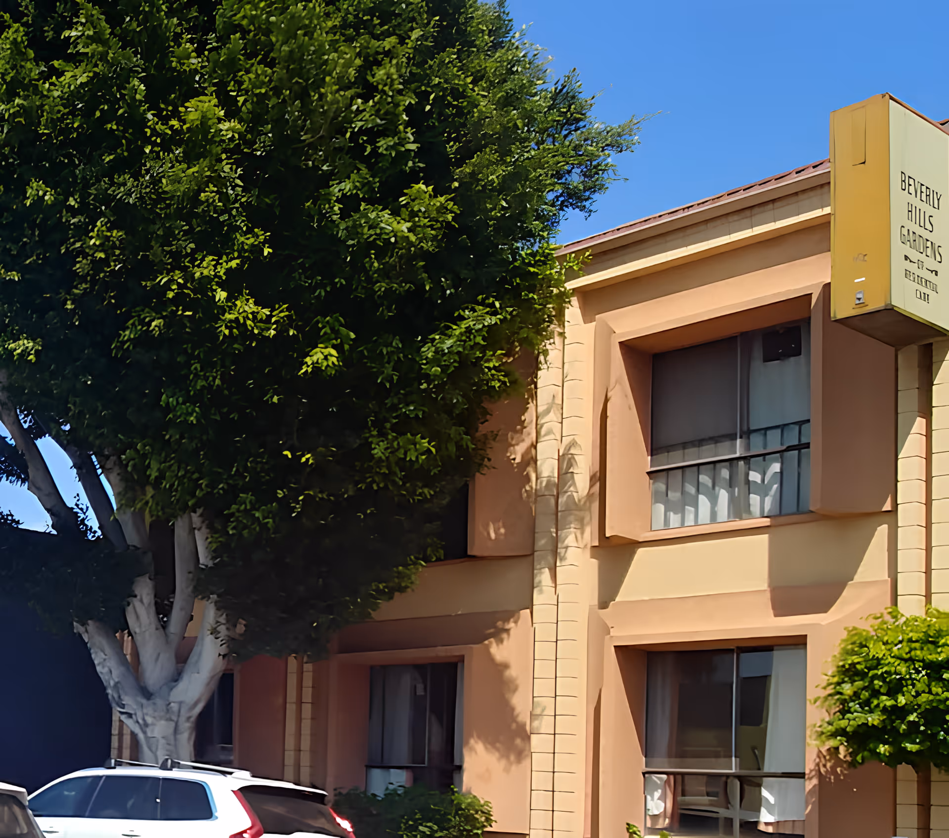 Exterior view of a two-story beige building with large windows partially covered by curtains, a large leafy tree in front, and a sign that reads 'Beverly Hills Gardens'. Several parked cars are visible in front of the building under a clear blue sky.