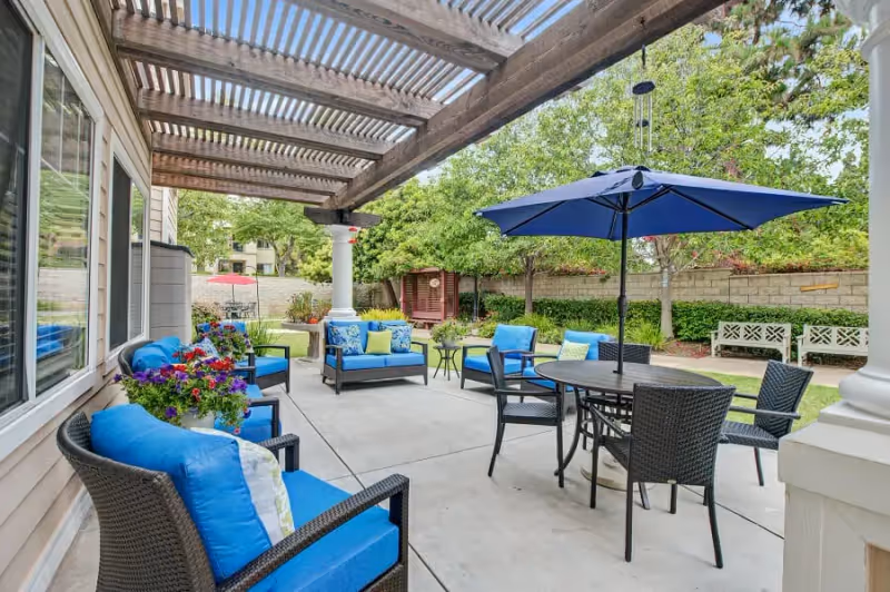 Outdoor patio area with blue cushioned wicker chairs, a round table with a blue umbrella, and a wooden pergola overhead. The patio overlooks a garden with trees, shrubs, a wooden bench, and a small gazebo in the background.