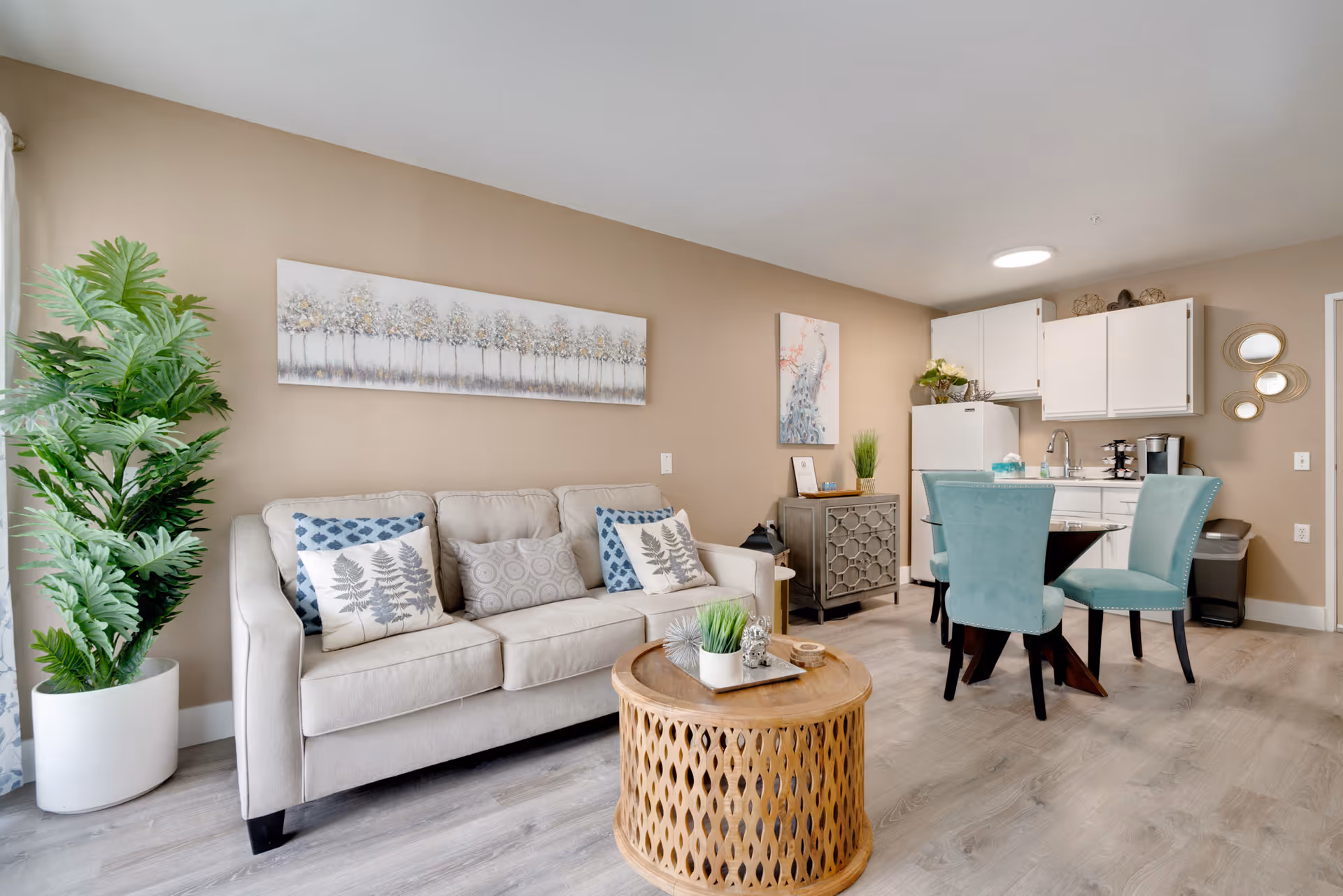 Bright open living area with a beige sofa, round wooden coffee table, large potted plant, and a small dining table with teal chairs beside a kitchenette.