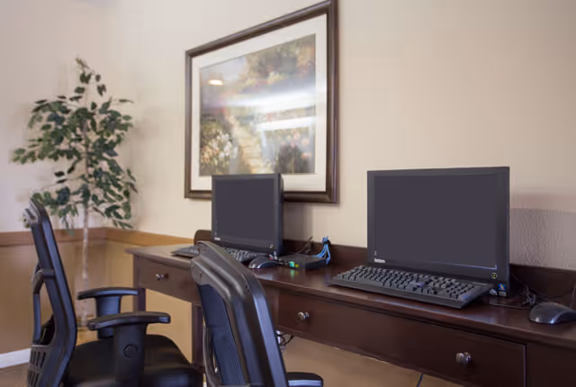 A workspace area in Bradfield Terrace featuring two desktop computers on a long wooden desk with two black office chairs. A framed painting of a garden scene hangs on the beige wall above the desk, and a potted plant is placed in the corner of the room.