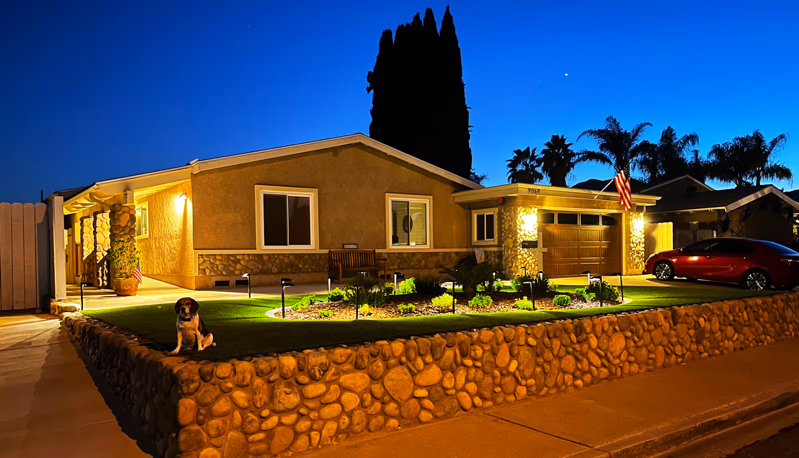 A single-story house at dusk with exterior lights on, a stone wall surrounding a well-maintained front yard with plants and a small dog sitting on the grass. There is an American flag near the garage and a red car parked in the driveway.