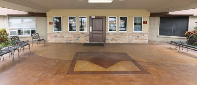 Covered entrance area of a building with a wooden door in the center, flanked by windows. The floor has a decorative pattern, and there are metal benches on both sides near the walls. Some plants are visible on the edges.