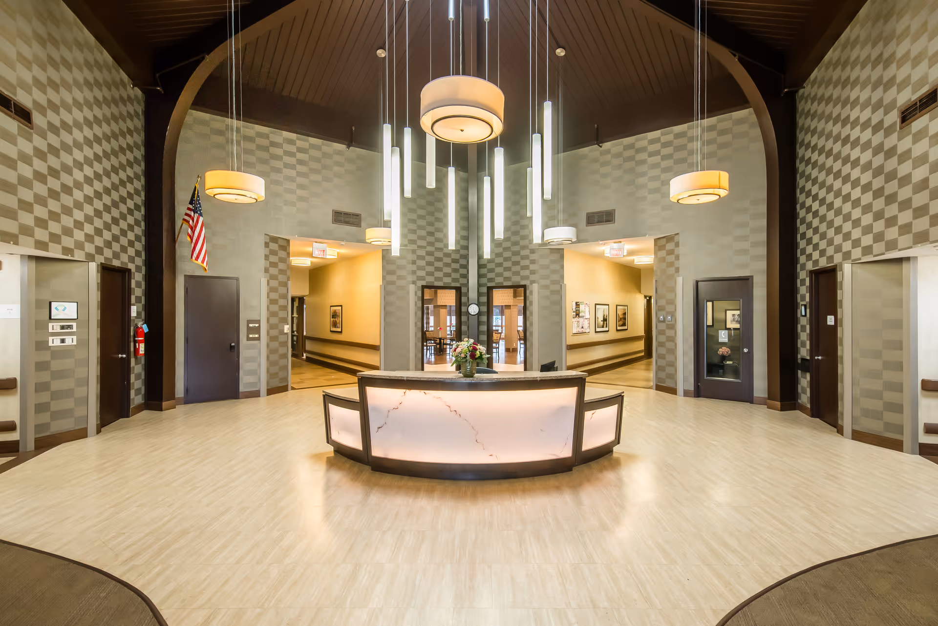 Spacious and well-lit reception area with a modern circular front desk illuminated from within, hanging pendant lights, checkered patterned walls, and multiple doorways leading to other parts of the facility. An American flag is displayed on the left side wall.