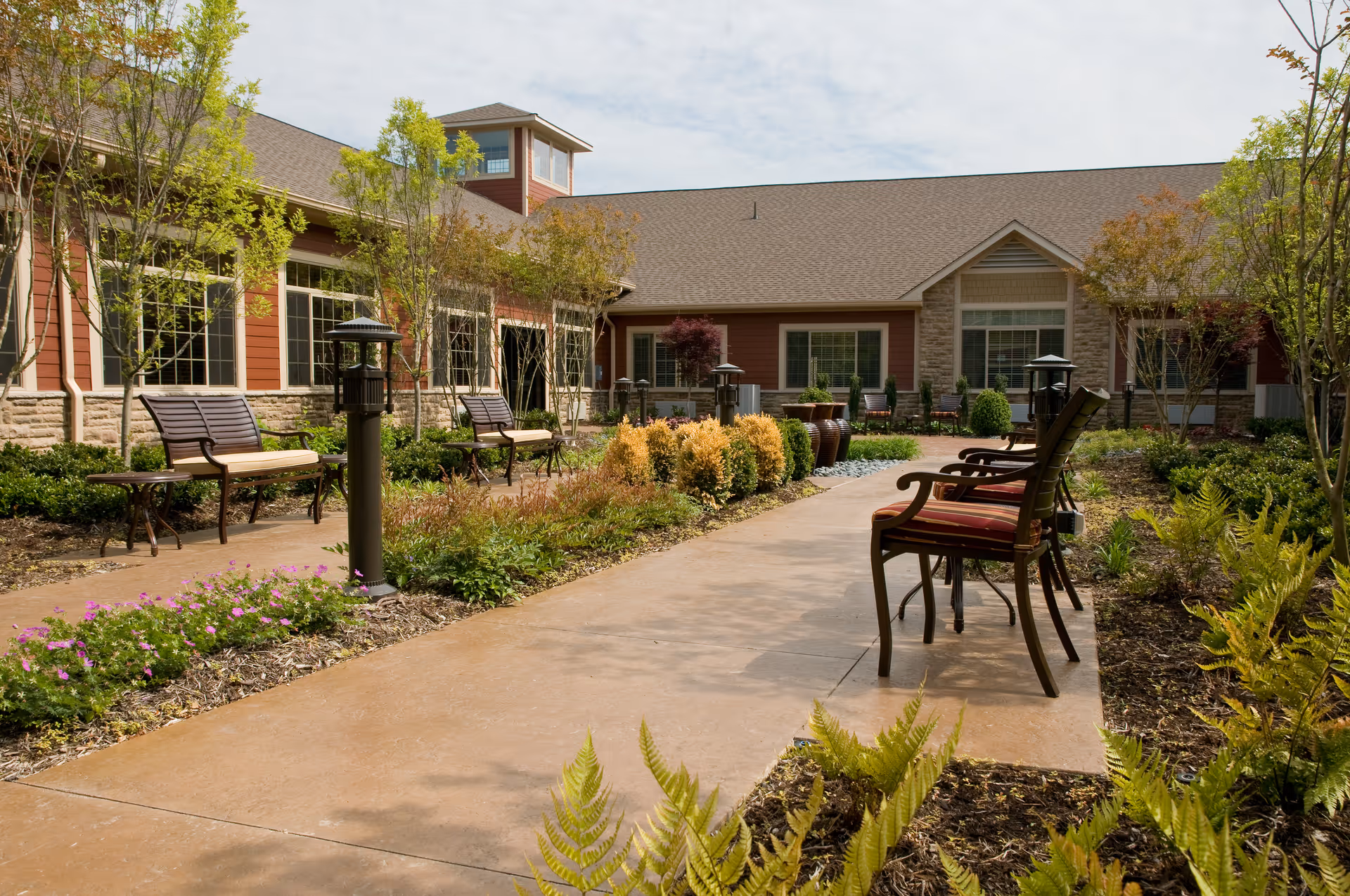 Sunlit courtyard with a paved walkway, benches and chairs surrounded by landscaped plantings in front of a single-story facility building.