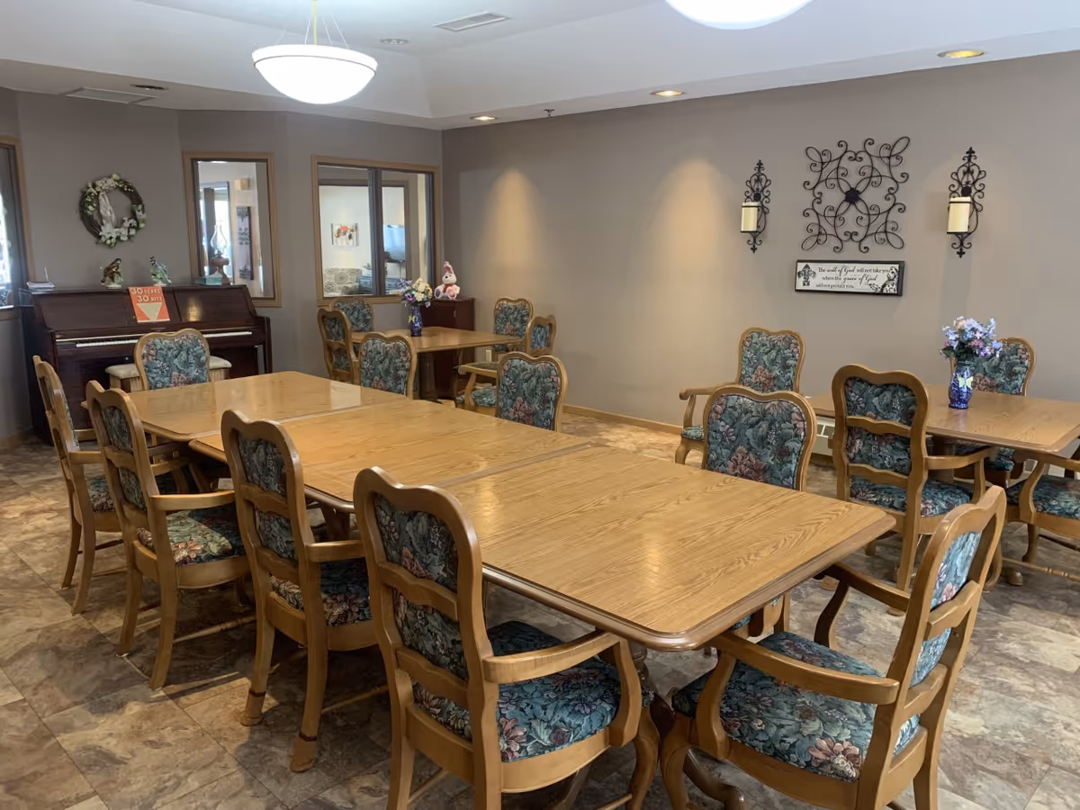 A dining room with multiple wooden tables and floral upholstered chairs arranged around them. There is a piano against the wall with a wreath and decorative items on top. The walls are beige with decorative wall hangings and a framed quote. The floor has a tile pattern, and the room is well-lit with a ceiling light fixture.