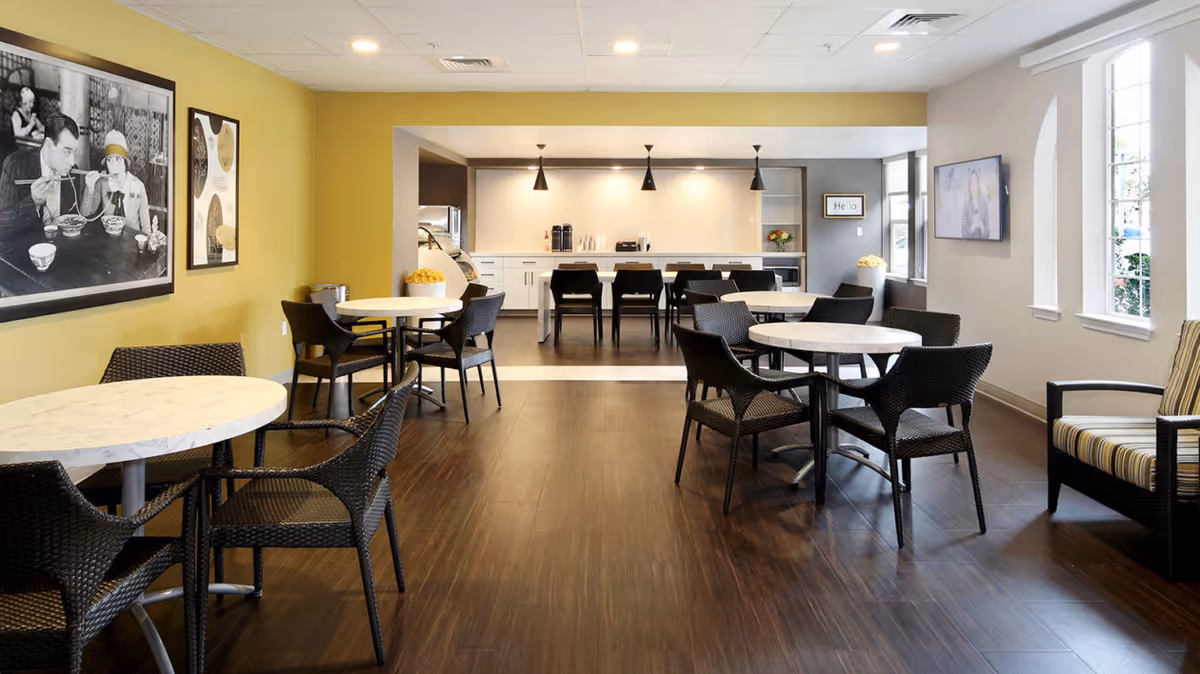 A bright and modern dining area with round white marble tables and black wicker chairs. The room features a yellow accent wall with framed black and white photos, dark wood flooring, and a kitchen area in the background with pendant lights and a counter with chairs. Large windows on the right side allow natural light to fill the space.