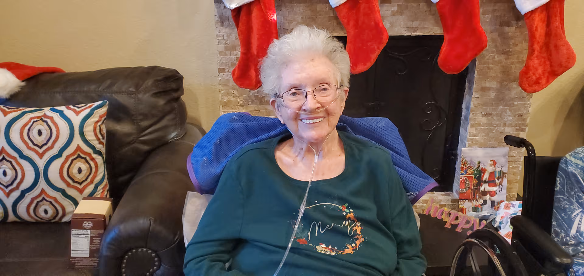 An elderly woman with white hair and glasses is sitting in a wheelchair in a cozy living room. She is smiling and wearing a green shirt with a festive design. Behind her is a fireplace decorated with red Christmas stockings and holiday cards. To her left is a dark brown leather couch with a colorful patterned pillow.