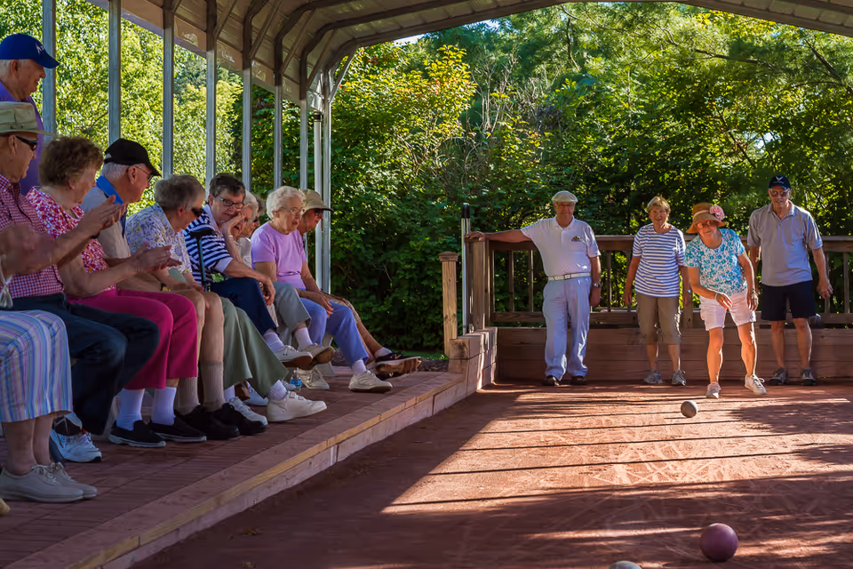 A group of elderly people at Heath Village Retirement Community enjoying a game of bocce ball under a covered outdoor area. Some participants are actively playing while others sit on benches watching and clapping. The scene is surrounded by lush green trees and natural light.