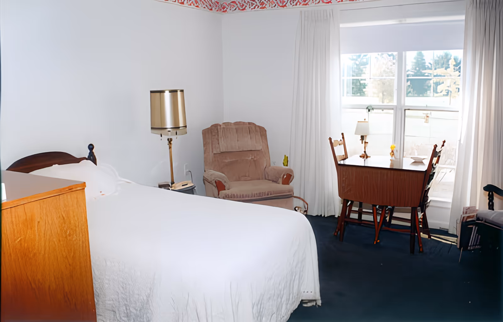 A bedroom in Elmwood Assisted Living & Skilled Nursing of Fremont featuring a bed with white bedding, a wooden dresser, a beige upholstered armchair, a floor lamp, and a small wooden dining table with two chairs near a large window with white curtains.