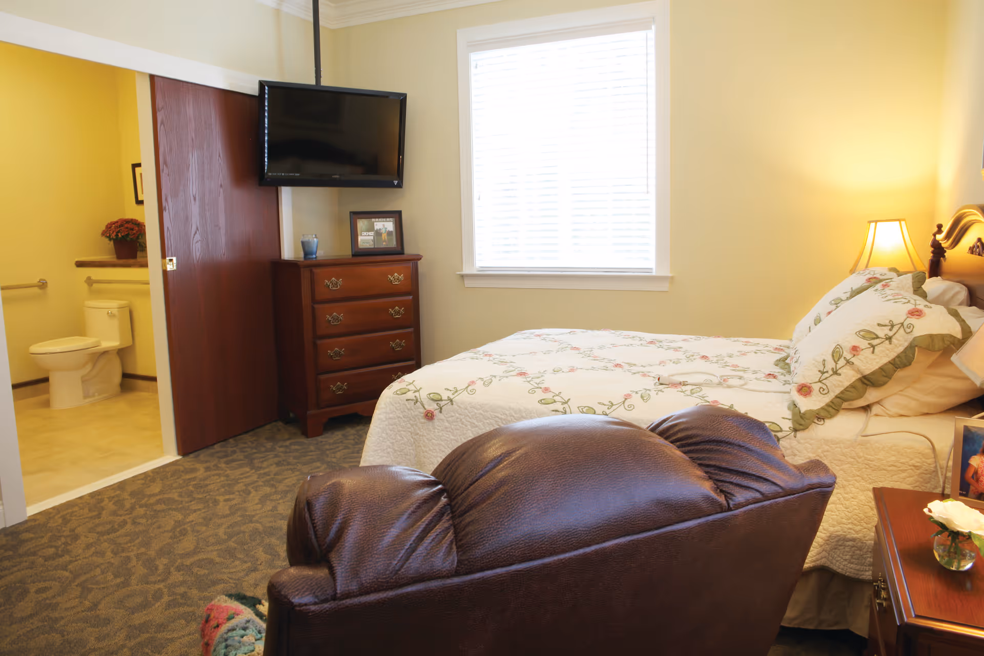 A cozy bedroom with a floral quilted bedspread, a wooden dresser with a TV mounted above it, a leather armchair, and a window with blinds. The room has soft yellow walls and carpeted floor. An open door reveals a bathroom with a toilet and a potted plant on the shelf.