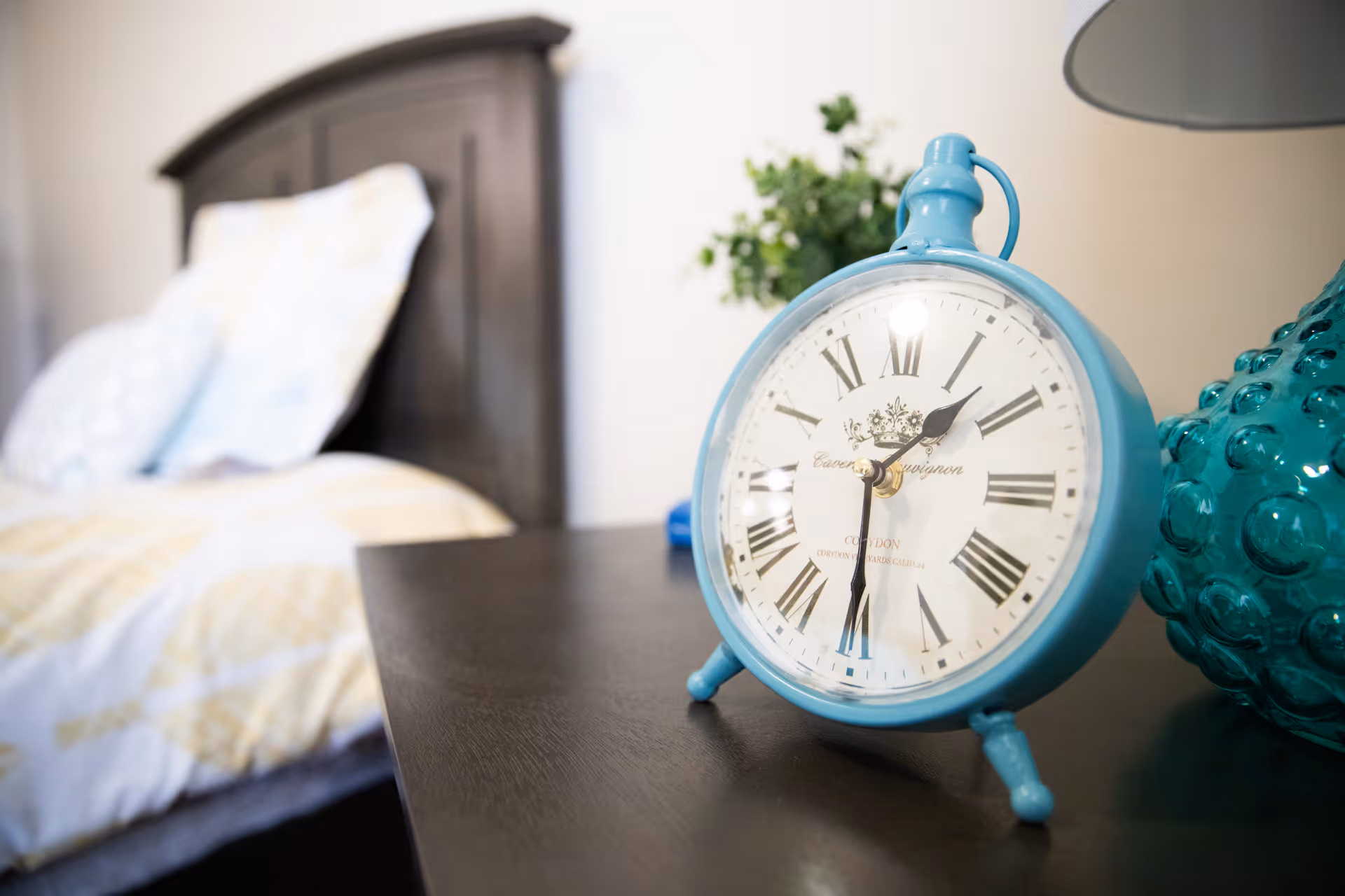 A bedside table in a bedroom featuring a turquoise vintage alarm clock, lamp, and a blurred bed in the background.