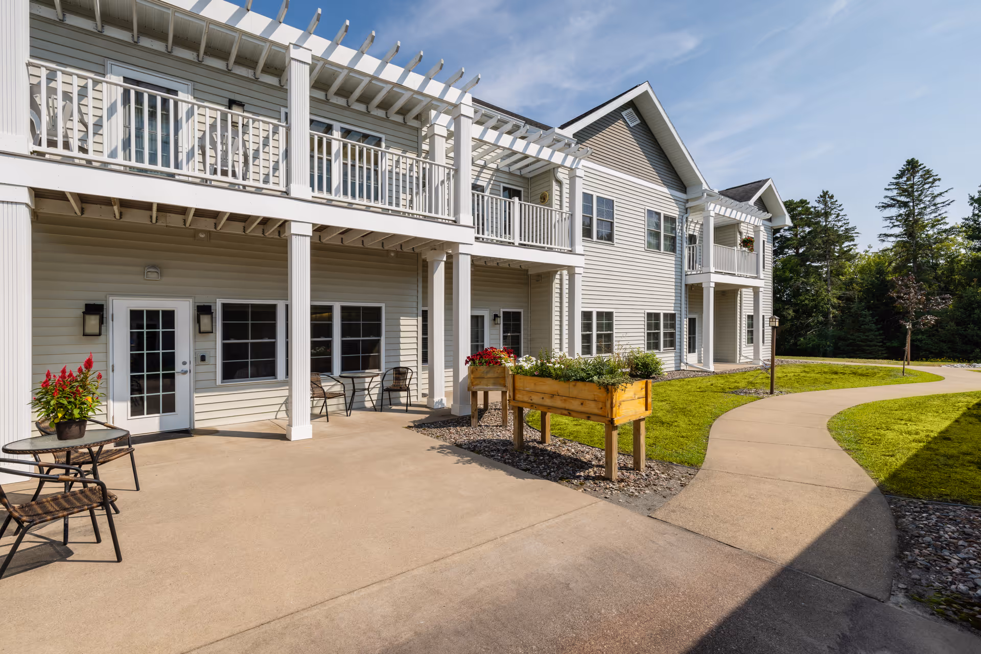 Exterior view of a two-story senior living facility building with beige siding, white railings, and balconies. There is a paved walkway curving through a green lawn with trees in the background. Outdoor seating with tables and chairs is visible on the concrete patio area, along with raised garden beds containing plants.