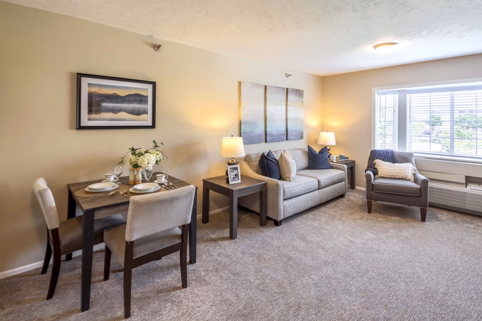 Bright living room with a sofa, armchair, side tables and a small dining table set near a large window.
