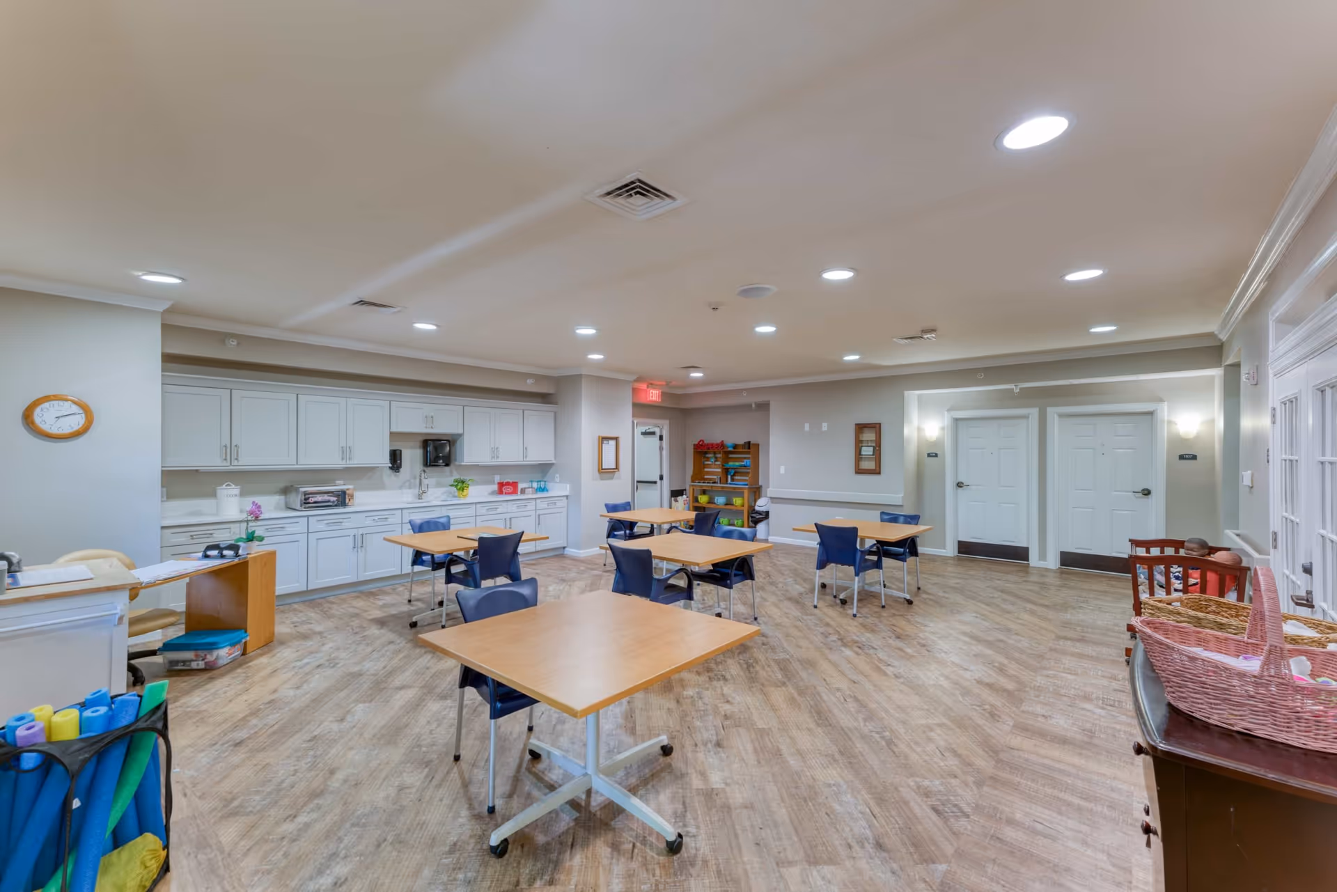 A spacious, well-lit room with light wood flooring and white walls, featuring several square tables with blue chairs arranged around them. Along one wall, there is a long counter with white cabinets, a microwave, and a toaster oven. The room has recessed ceiling lights, a clock on the wall, and a few shelves with books and games. There are multiple doors visible, and a basket with pink lining is on a dark wooden cabinet in the foreground.