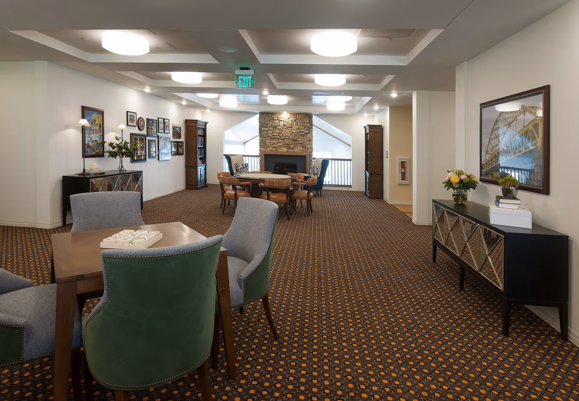 A spacious, well-lit common area with patterned carpet, several tables and chairs, a stone fireplace at the far end, framed pictures on the walls, and decorative furniture including a sideboard with flowers and books.