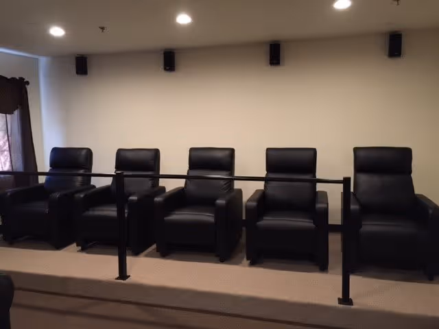 A row of five black leather recliner chairs arranged on a raised platform with a black metal railing in front, set against a plain beige wall with three black speakers mounted near the ceiling.