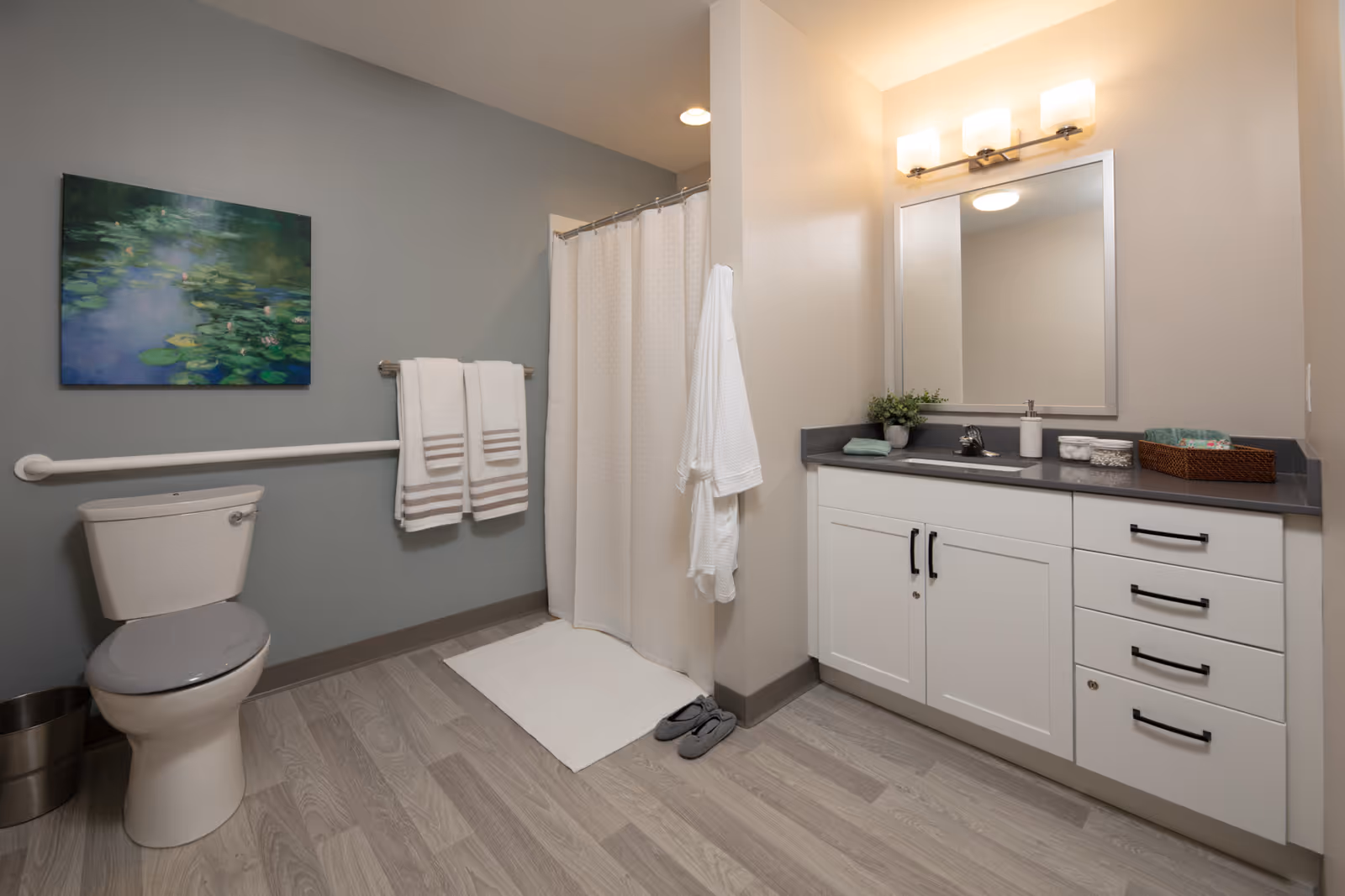 A clean and modern bathroom featuring a toilet with a gray seat, a white towel rack holding two white towels with gray stripes, a shower area with a white curtain, a white bathrobe hanging on the wall, a white bath mat, gray slippers on the floor, and a vanity with a gray countertop, white cabinets, a large mirror, and a light fixture above. A green plant and bathroom essentials are placed on the countertop. The walls are painted in light gray and beige tones, and a painting of water lilies hangs above the toilet.