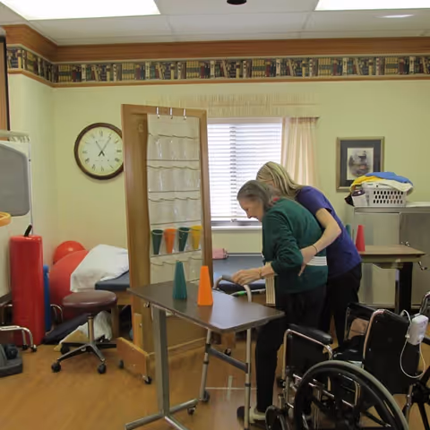 An elderly woman receiving assistance from a caregiver in a therapy or rehabilitation room. The elderly woman is standing with support near a table with colored cones, and a wheelchair is nearby. The room has a clock on the wall, a window with blinds and curtains, and various therapy equipment.
