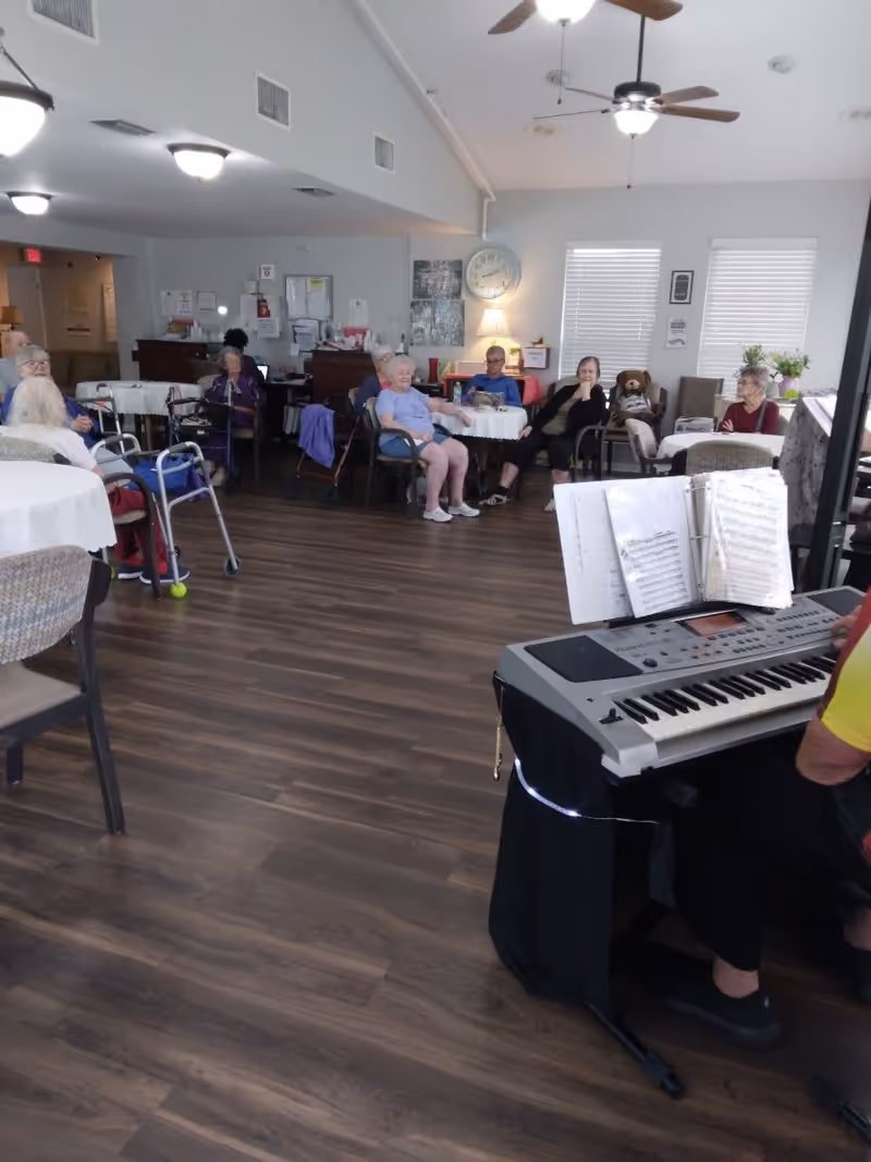 A group of elderly people sitting around tables in a common room with wooden flooring. A person is playing an electronic keyboard with sheet music in the foreground. The room has ceiling fans, windows with blinds, and various decorations including a clock and a teddy bear on a chair.