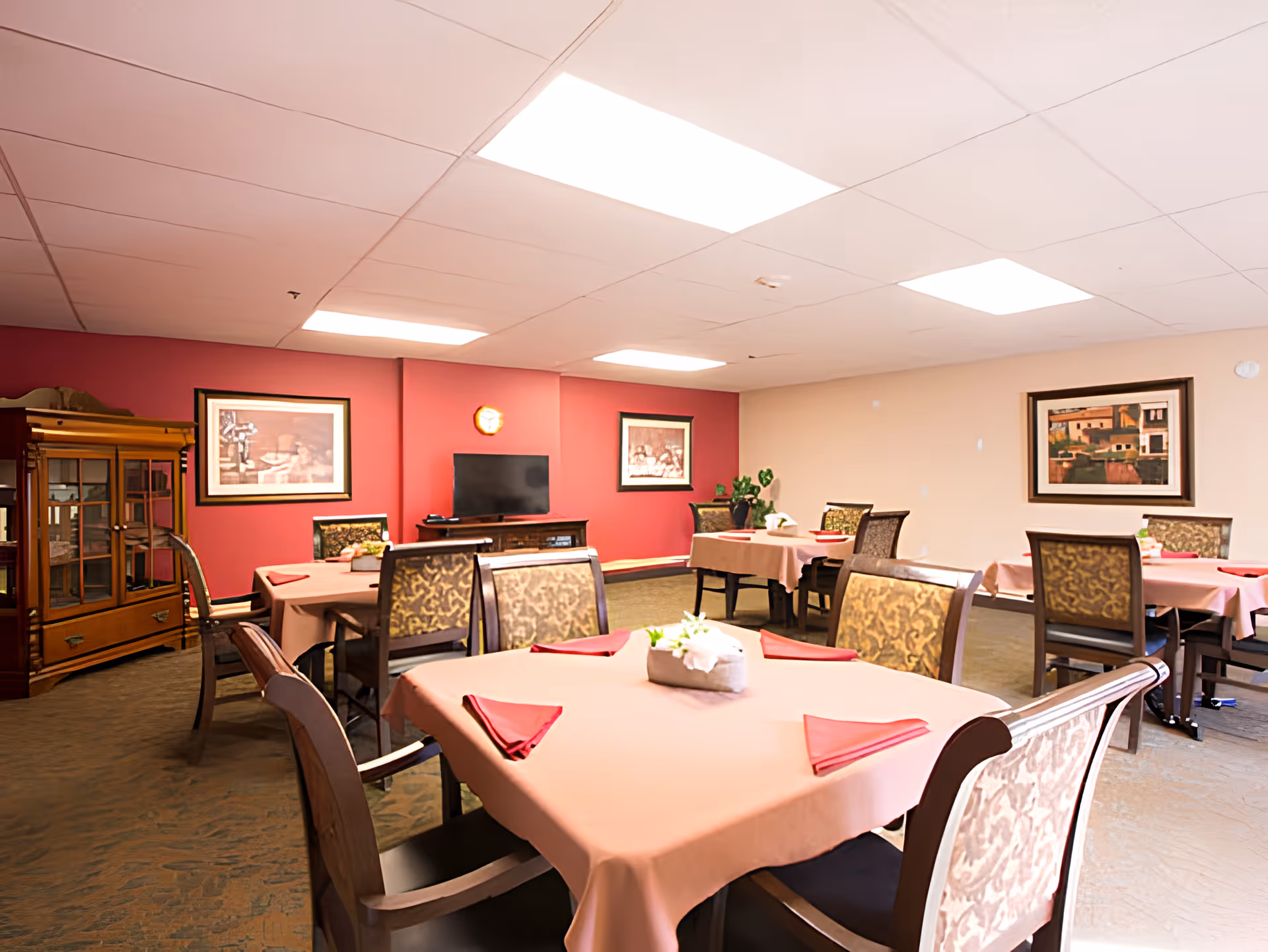 A dining room in a senior living facility with several tables covered in light pink tablecloths and red napkins. Each table has a small floral centerpiece. The room has patterned chairs, a red accent wall with framed artwork, a TV on a wooden stand, and a wooden cabinet. The ceiling has recessed lighting panels.