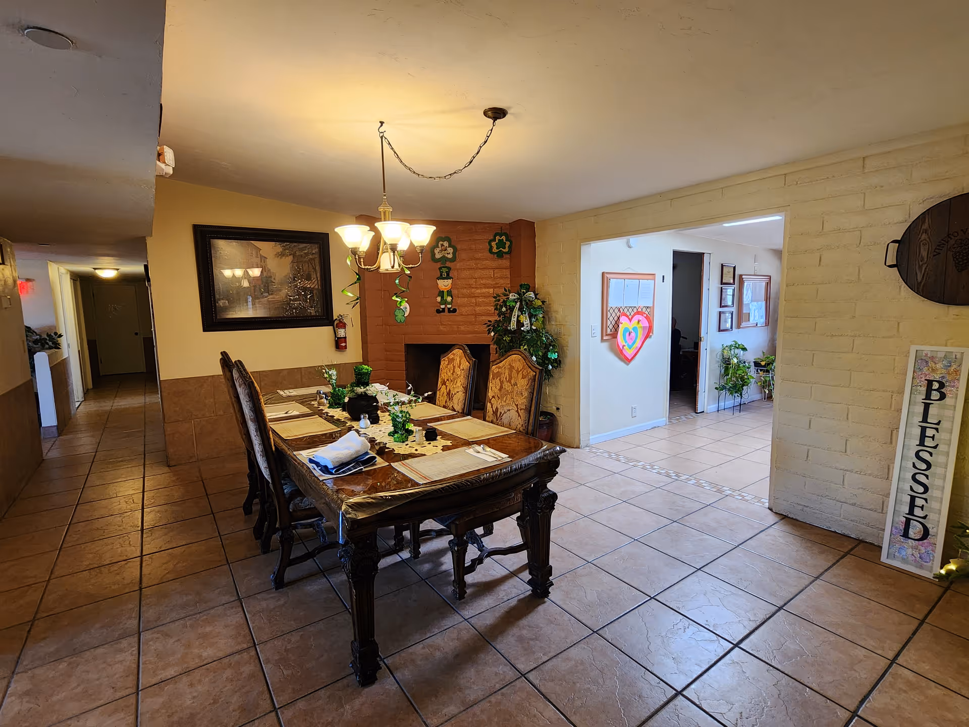 A dining area in an assisted living facility with a wooden dining table set for six, decorated with green shamrocks and a leprechaun figure on the brick fireplace. The room has tiled floors, a chandelier overhead, and a hallway leading to other rooms. A decorative sign with the word 'BLESSED' is visible on the right wall.
