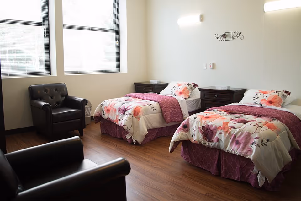 A bright room with two single beds featuring floral bedspreads and purple blankets. Each bed has a dark wooden nightstand beside it. There are two black leather armchairs in the room, and two large windows with blinds letting in natural light. The floor is wooden, and the walls are light-colored with minimal decoration.