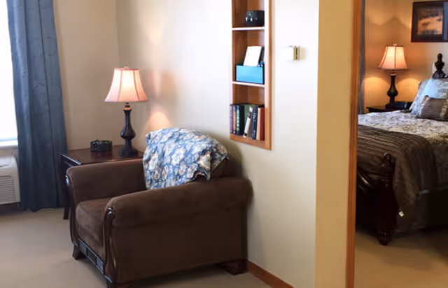 Cozy sitting area with a brown armchair, side table and lamp, built-in wall shelf, and a view into a bedroom with a bed and bedside lamp.