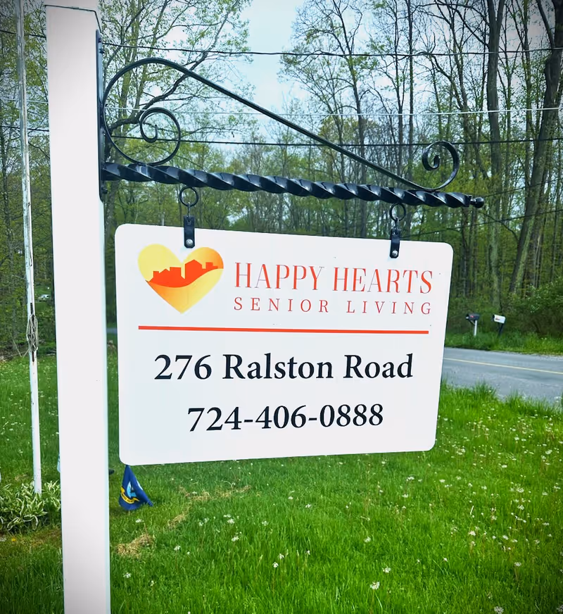 A white hanging sign with a black decorative metal bracket displaying the logo and contact information for Happy Hearts Senior Living. The sign features a heart-shaped logo with a cityscape inside, the text 'HAPPY HEARTS SENIOR LIVING', the address '276 Ralston Road', and the phone number '724-406-0888'. The sign is set outdoors with green grass and trees in the background.