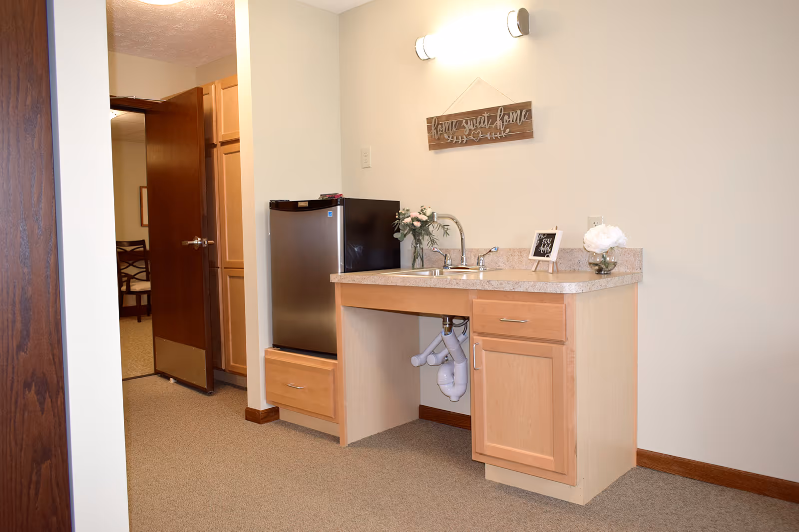 Compact kitchenette with a countertop sink, light wood cabinets, a mini refrigerator, and wall decor.