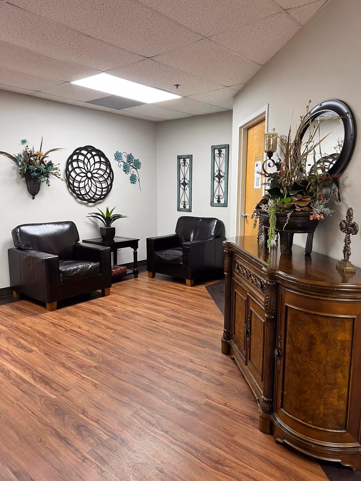 A cozy seating area with two black leather armchairs separated by a small black side table with a potted plant. The walls are decorated with metal wall art and floral arrangements. To the right, there is a wooden cabinet with intricate carvings, topped with a large floral arrangement and a round mirror above it. The floor is wood laminate, and the ceiling has fluorescent lighting.