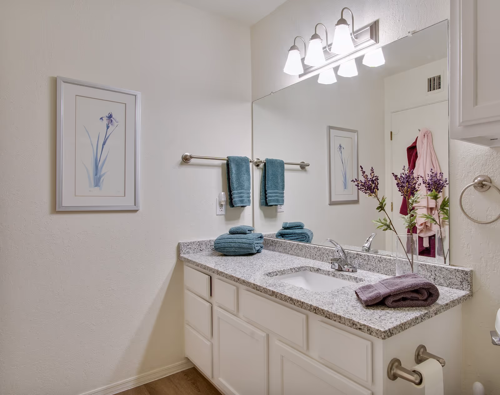 Bathroom vanity with granite countertop, sink, large mirror, teal towels, lavender flowers, and framed wall art.
