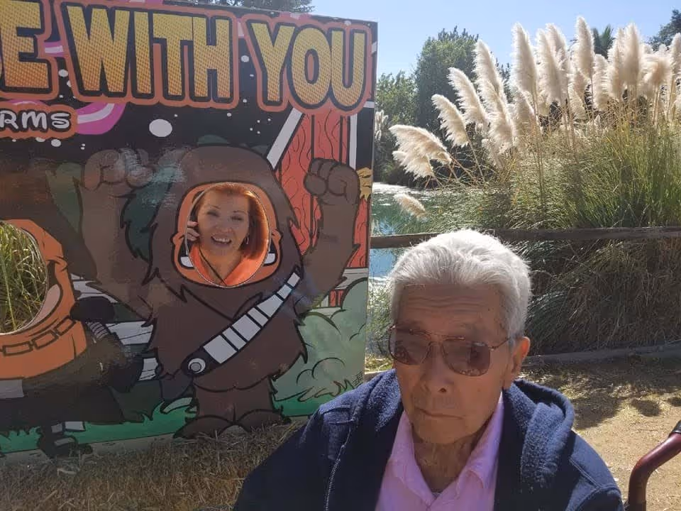 An elderly man wearing sunglasses and a blue jacket sits outdoors near tall ornamental grasses. Behind him is a colorful cutout board with cartoon characters, one of which has a face cutout where a smiling woman is posing. The board has large text partially visible that reads 'WITH YOU'.