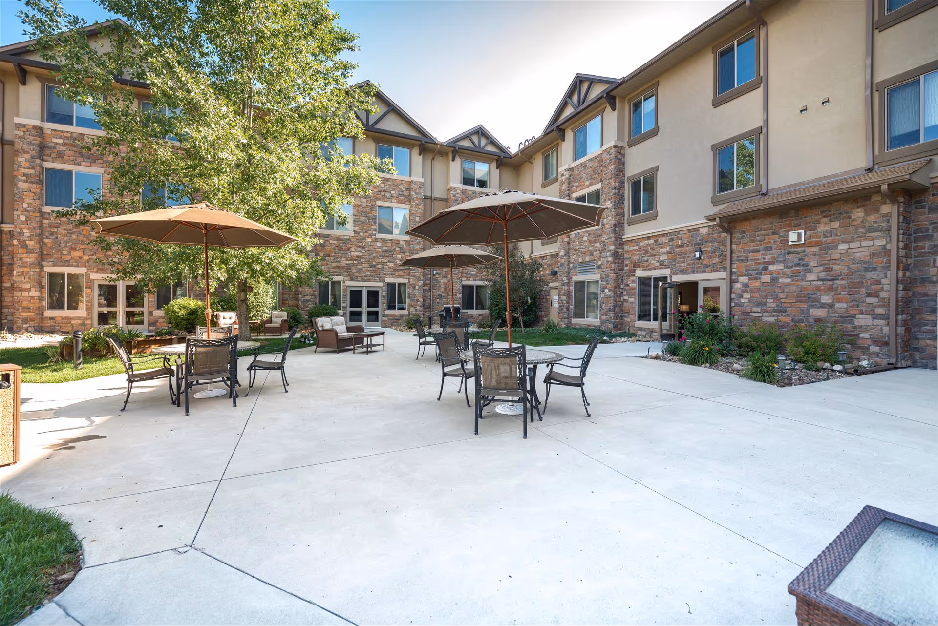 Outdoor courtyard area of a senior living facility with several round tables and chairs under large umbrellas, surrounded by a three-story building with stone and stucco exterior walls, windows, and some greenery including a large tree and bushes.