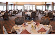 A spacious dining room with multiple round tables covered with white tablecloths, each set with plates, glasses, silverware, and folded maroon napkins. The room has large windows allowing natural light to fill the space, and upholstered chairs surround each table.