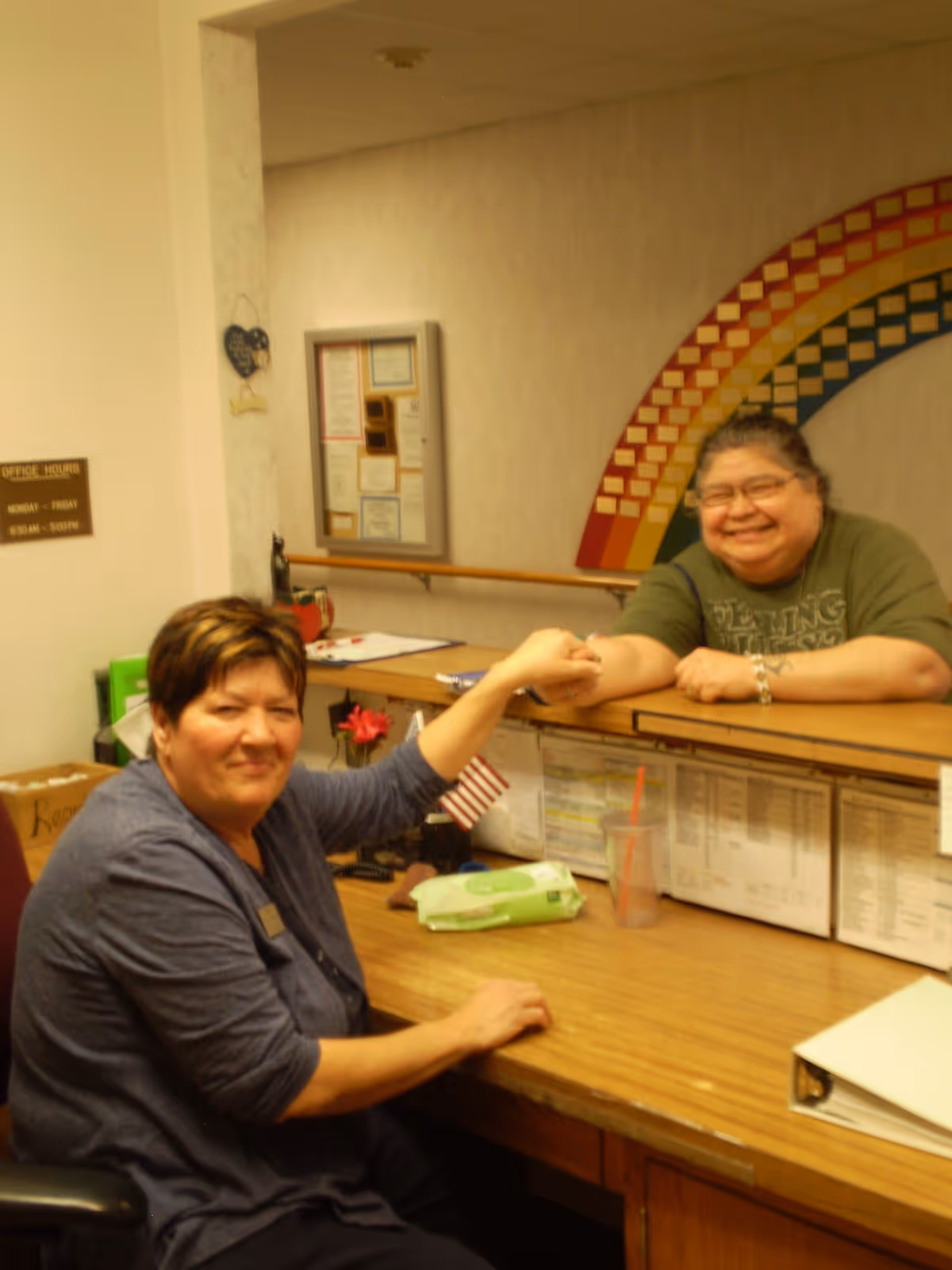 Two women smiling and holding hands across a reception desk in an office area. One woman is seated behind the desk, and the other is leaning on the counter. The background features a colorful rainbow wall decoration and a bulletin board with papers.