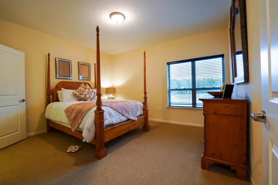 A cozy bedroom with a wooden four-poster bed dressed in white bedding and a pink throw blanket. There are two framed artworks on the wall above the bed, a bedside lamp on a small table, a wooden dresser with a mirror, and a window with blinds letting in natural light. A pair of slippers is placed on the carpeted floor near the bed.