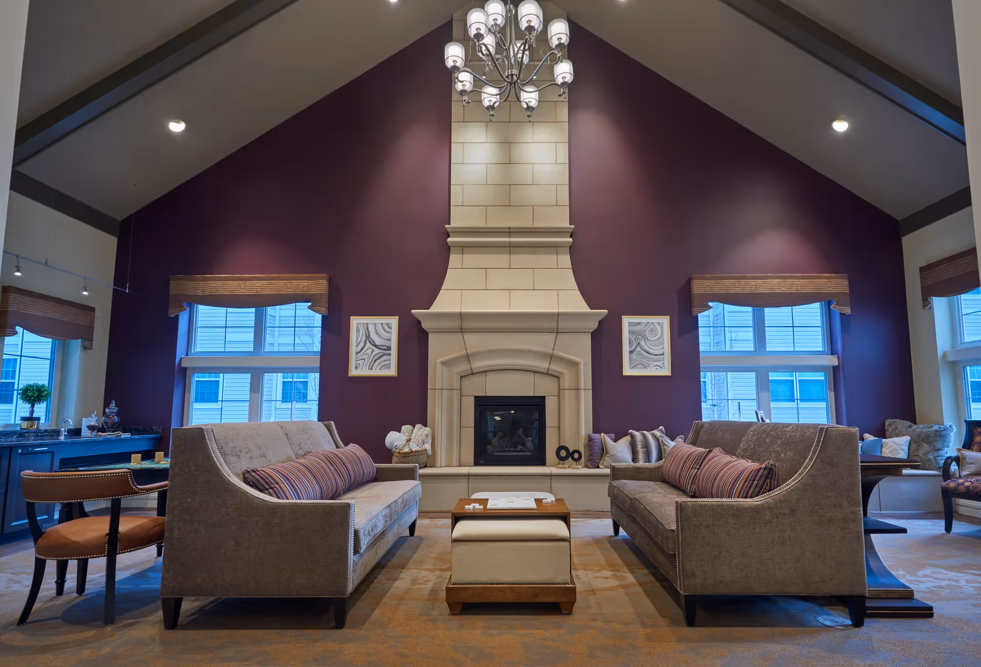 A spacious and elegant living room with two gray sofas facing each other across a small ottoman and wooden coffee table. The room features a tall beige stone fireplace centered on a deep purple accent wall, flanked by two windows with wooden valances. A chandelier hangs from the vaulted ceiling, and additional seating and a desk area are visible on the sides.