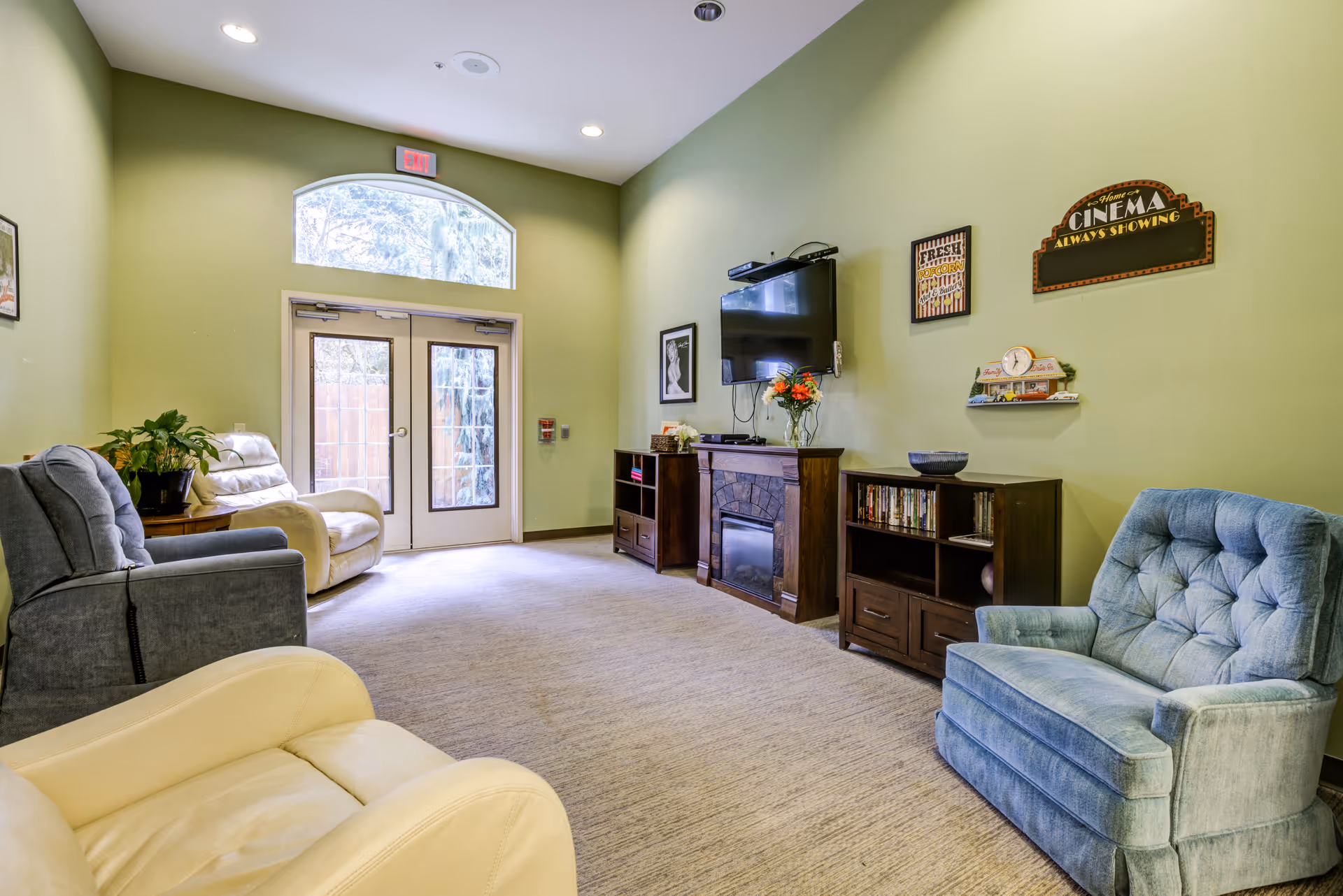 A cozy living room with light green walls, featuring several comfortable armchairs in beige and blue colors arranged around a TV mounted above a fireplace. There are two wooden cabinets with books and decorative items, a vase with flowers, and wall decorations including a sign that says 'Cinema Always Showing'. A large window above double glass doors lets in natural light.