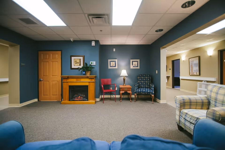 A cozy sitting area in a senior living facility with a blue wall featuring a wooden electric fireplace, a red chair, a small wooden side table with a lamp, and a patterned armchair. The room has carpeted flooring, framed artwork on the walls, and a hallway leading to other rooms. Additional seating includes a plaid armchair and a blue couch partially visible in the foreground.